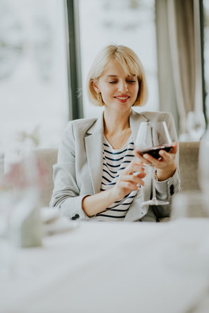 A woman with short blonde hair smiles as she examines a glass of red wine. She sits at a well-set table in a modern restaurant, surrounded by soft lighting and elegant decor.の写真素材