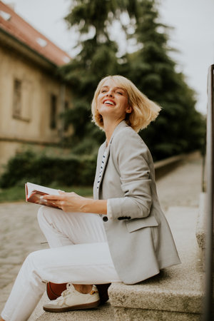 A joyful woman sits on steps, reading a book. Her light gray blazer and white pants complement her carefree smile. The background features lush greenery and historic architecture.の写真素材