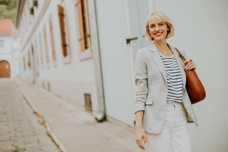 A cheerful woman strolls along a quaint street in a charming European town. She wears a striped shirt and light blazer, carrying a small bag. Sunlight adds warmth to the scene.の写真素材