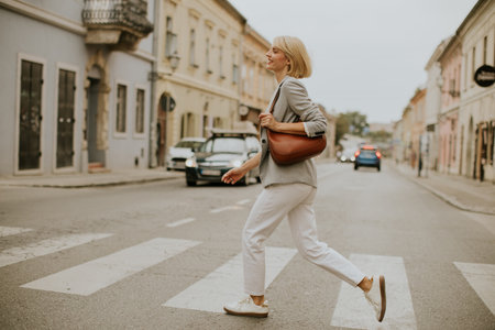 A woman walks confidently across a zebra crossing in a city. She wears a light gray blazer, white pants, and sneakers. Vehicles are parked nearby as she strides along the road.の写真素材