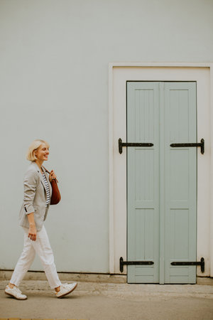 A woman strolls casually along a street, wearing a relaxed outfit with white pants and a gray blazer. She carries a brown bag while enjoying her day near a pastel blue door.の写真素材