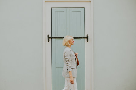 A woman with short blonde hair walks confidently next to a light blue door, holding a small bag. She...