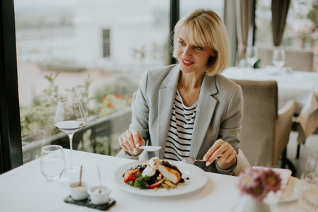 A woman in a stylish blazer sits at a table in a contemporary restaurant, savoring a gourmet dish. She smiles as she prepares to take a bite while enjoying the scenic view.の写真素材