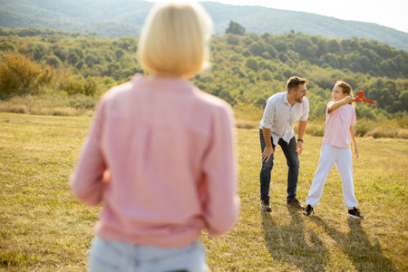 A family spends quality time outdoors on a sunny day in a grassy field. A girl prepares to throw a red object while her father watches with enthusiasm, and a woman observes nearby.の写真素材