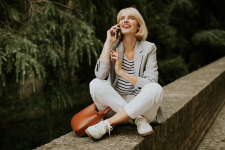 A cheerful woman sits on a stone wall in a green park, talking on her phone. She wears a light...