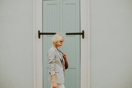A young woman with short blonde hair walks leisurely next to a light blue door. She carries a brown handbag and wears a stylish outfit, enjoying a sunny day in the coastal town.の写真素材