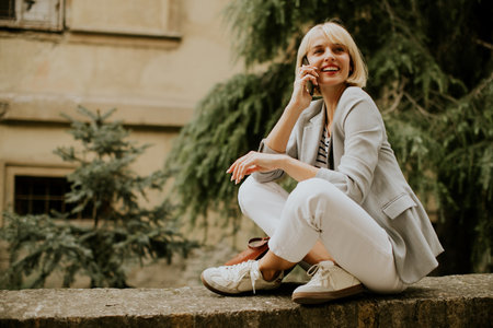 A young woman is sitting on a stone wall in an urban area, engaged in a phone conversation. She is smiling and wearing a light gray blazer with white pants and sneakers, enjoying the moment.の写真素材
