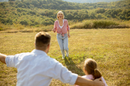 A woman in a pink blouse runs happily towards her family in a sunny field. The scene captures a warm moment filled with joy and connection against a background of green hills.の写真素材
