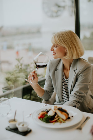 A woman in a gray blazer sips red wine while dining at an upscale restaurant. A plate of gourmet food is in front of her, and the city skyline is visible through the window.の写真素材