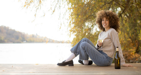 A young woman relaxes by the serene lake, basking in the warm autumn sunlight and vibrant surroundings.の写真素材