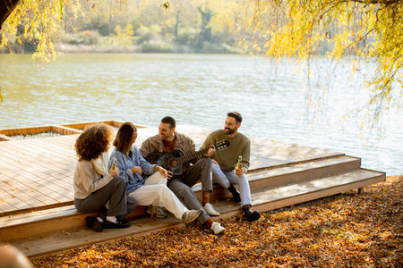 A group of four friends relax by a serene lake, sharing moments of joy, music, and laughter under the warm autumn sun.の写真素材