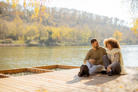 Two people sit on a wooden dock by a calm lake, sharing laughter and warmth on a bright autumn day surrounded by colorful trees.の写真素材