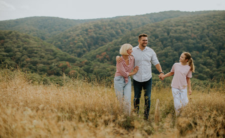 A family is walking hand in hand in a grassy field. They are smiling and enjoying the fresh air while surrounded by lush green mountains and golden sunlight in the early evening.の写真素材