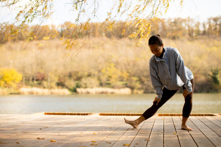 A Korean woman practices her stretching routine by a tranquil lake, surrounded by golden autumn leaves and a calming atmosphere.の写真素材