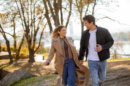 A young couple strolls hand in hand through a park filled with autumn trees. The sun shines brightly, casting a warm glow on their smiles and the vibrant leaves around them.の写真素材