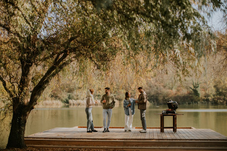 Friends gather around a grill on a wooden dock by a tranquil lake, sharing laughter and delicious food, surrounded by autumn colors.の写真素材