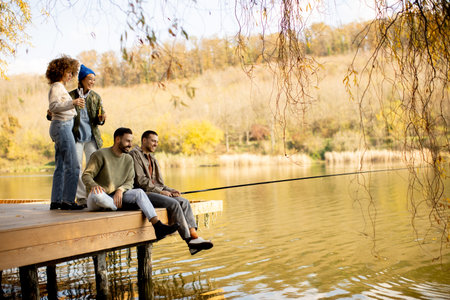 A group of friends relaxes on a dock by a serene lake, engaging in fishing. The colorful autumn...