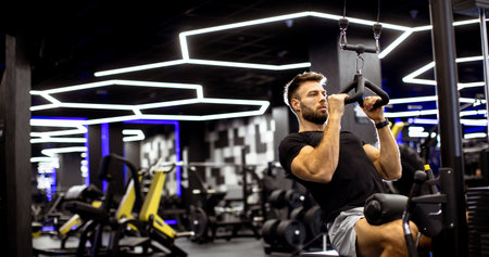 A fit man performs pull-ups on a specialized machine in a contemporary gym. The setting features...