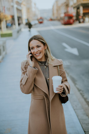 A woman smiles brightly while speaking on her phone and holding a coffee cup. She is dressed warmly in a coat, walking on a busy street lined with buildings and traffic in the background.の写真素材