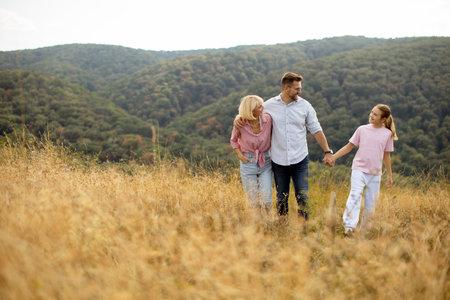 Family enjoys a relaxing walk hand in hand through a golden field on a sunny day. The hills in the background feature vibrant autumn colors.の写真素材