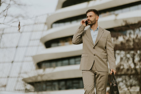 A suited man strolls along a modern building while engaged in a phone conversation, exuding professionalism and poise.の写真素材