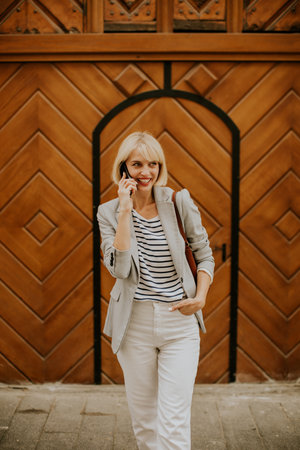 A young woman with blonde hair dressed in a striped shirt and blazer stands outside near a wooden door, smiling as she talks on her phone. Her stylish look suggests confidence and joy.の写真素材