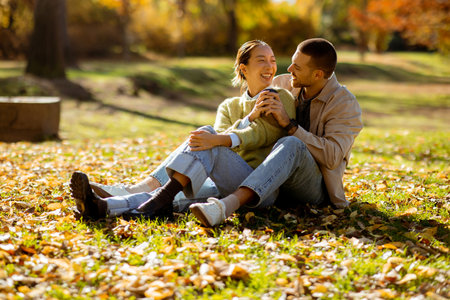 Under the warm sunlight, a Korean woman and a Caucasian man share a joyful moment sitting amidst colorful autumn leaves, sipping drinks together.の写真素材