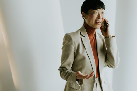 A woman in a beige suit has an animated conversation on her phone in a contemporary office. Her expression shows engagement as she interacts with the caller.の写真素材