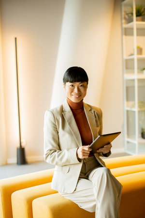 A confident business professional sits on a yellow couch in a modern office. She is smiling and using a tablet while surrounded by a clean, stylish interior with soft lighting.の写真素材