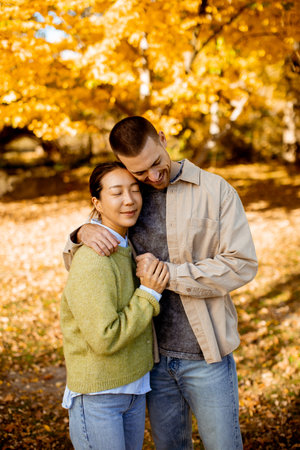 A Korean woman in a green sweater and a Caucasian man lean close together in a beautiful autumn park. The leaves shimmer in gold.の写真素材