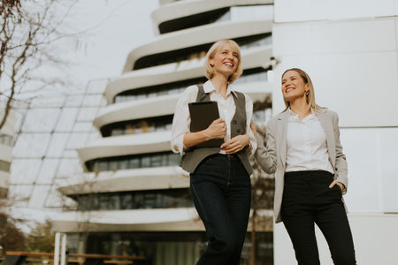 Two women in business attire smile and chat as they walk outside a contemporary building. They carry a tablet and exude a positive energy, showcasing professionalism and collaboration.の写真素材