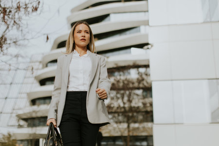 A businesswoman in formal attire is walking purposefully outside a contemporary building. She carries a bag and appears focused on her destination. It is a clear day with bright lighting.の写真素材