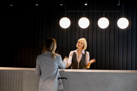 A woman with short blonde hair is welcoming a visitor at a sleek reception desk. The setting features modern lighting and a stylish design, creating a professional atmosphere.の写真素材