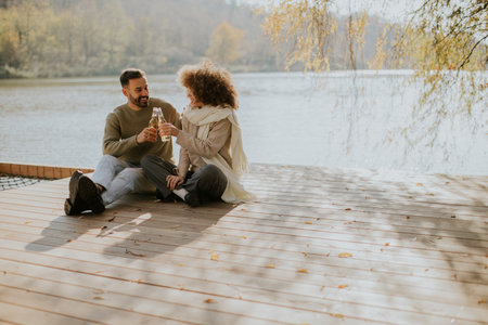 Two friends sit on a wooden dock by the lake, sharing laughter and drinks as the sun sets, surrounded by autumn colors.の写真素材