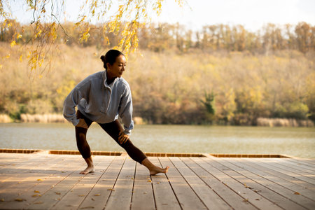 A Korean woman performs elegant dance poses on a wooden deck by a tranquil lake surrounded by vibrant autumn trees.の写真素材