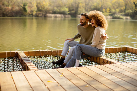 Smiling couple sits together on a wooden platform at a tranquil lake, surrounded by colorful autumn leaves.の写真素材