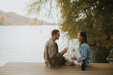 A couple enjoys each other's company on a lakeside deck, sipping drinks and laughing under the sun,...