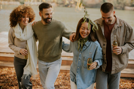 Four friends stroll by a serene lake, enjoying warm laughter and refreshing drinks, surrounded by autumn leaves and sunshine.の写真素材