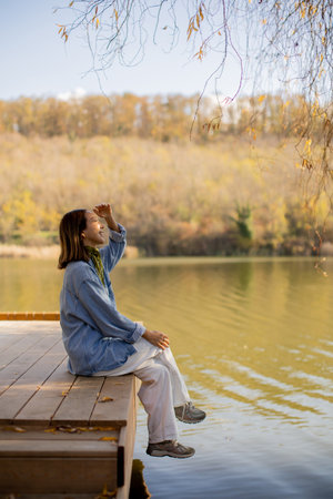 A woman sits on a wooden dock, gazing thoughtfully at the calm lake surrounded by vibrant autumn trees.の写真素材