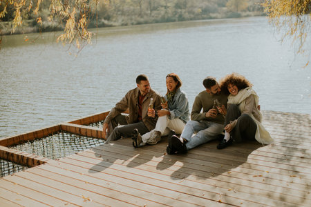 Group of friends relax by the water, sharing joyful moments and drinks under sunlight. Nature surrounds them, creating a serene atmosphere.の写真素材