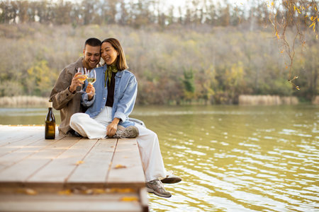 An enchanting scene unfolds as a couple sits on a wooden dock, sharing smiles and raising glasses of wine.の写真素材