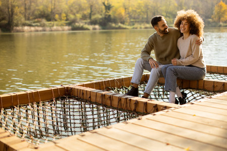 A couple sits closely on a wooden platform by the lake, surrounded by vibrant fall foliage, sharing joyful laughter and smiles.の写真素材