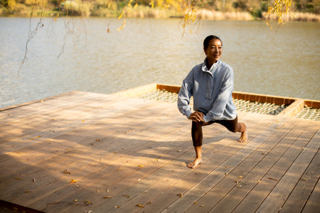 A woman in a cozy sweatshirt performs lunges on a wooden deck by the calm lake, enjoying the sunny weather and beautiful scenery.の写真素材