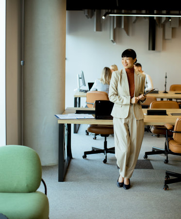 In a contemporary office, a business professional stands with a clipboard. Colleagues work at desks in the background. The environment showcases a blend of style and productivity.の写真素材