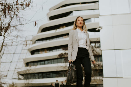 A businesswoman walks with purpose in a professional outfit, carrying a handbag, in front of a striking modern building. The scene captures urban life and ambition in a bustling city.の写真素材