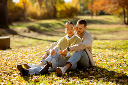 Two people share a joyful moment sitting together on the grass in a park. Bright autumn leaves create a beautiful backdrop.の写真素材