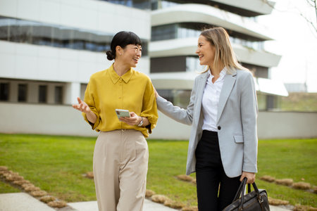 Two women walk together in a green area outside a contemporary building. They are laughing and discussing something while one holds a phone. The weather looks pleasant.の写真素材