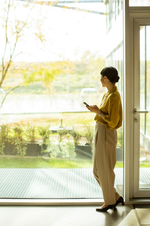 A woman in a yellow blouse and beige pants stands by a large glass window. She checks her phone while looking outside at greenery. The sunlight streams in, creating a bright atmosphere.の写真素材