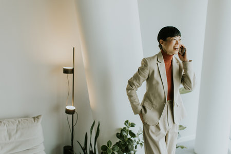 A woman dressed in a beige suit enjoys a phone conversation while standing in a stylish room filled with greenery. Bright light highlights her joyful expression and modern surroundings.の写真素材