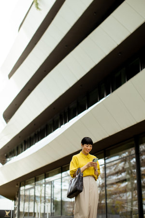 A woman dressed in a bright yellow blouse stands outside a contemporary building, smiling while looking at her smartphone. The sleek, curved design of the architecture surrounds her.の写真素材